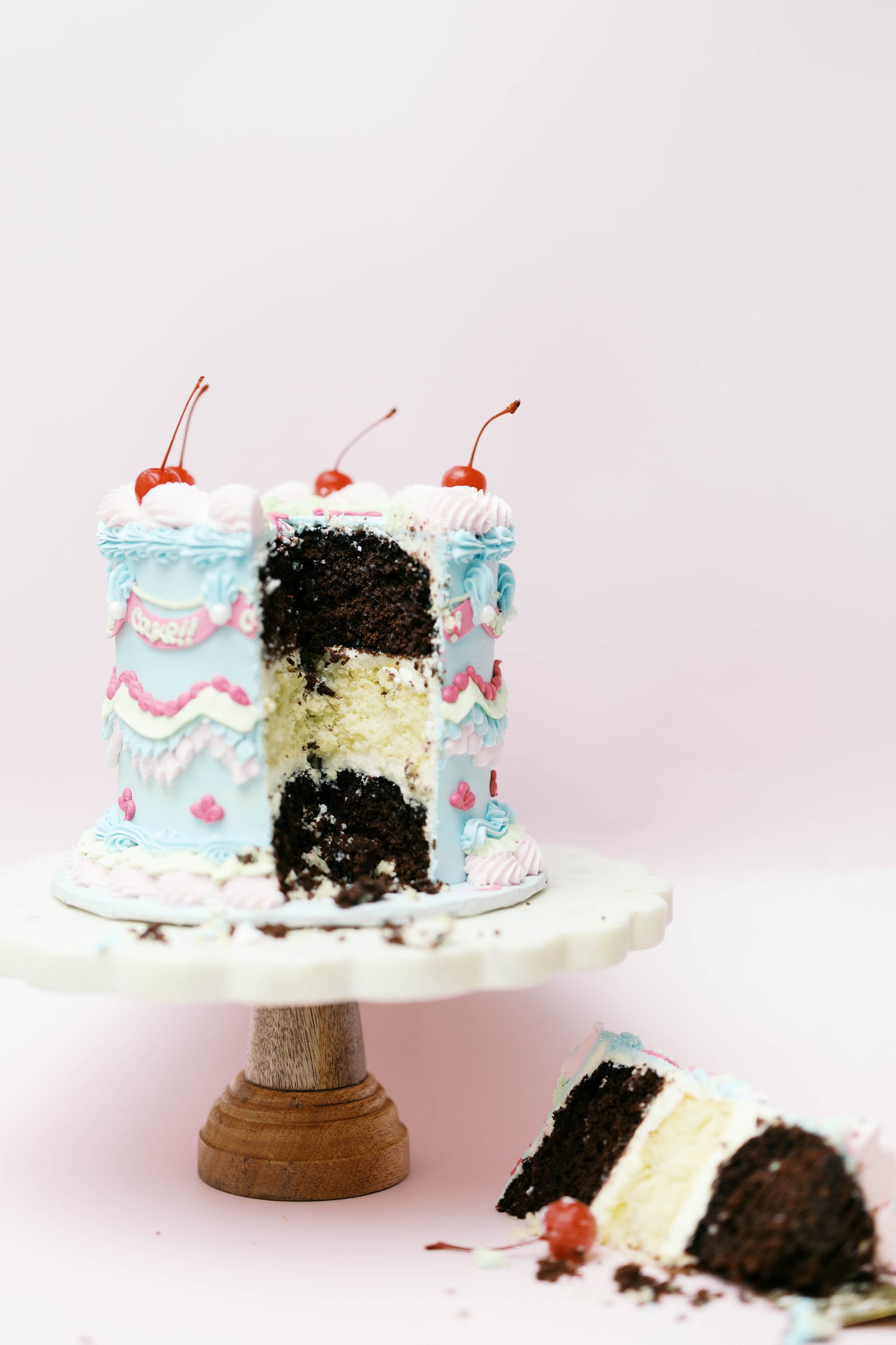 Small cake with white frosting, chocolate layers, and cherries on a pink background
