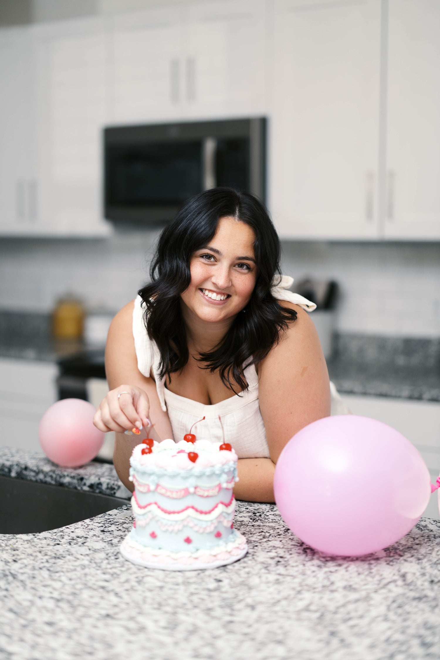 Woman in a kitchen with a small cake and pink balloons