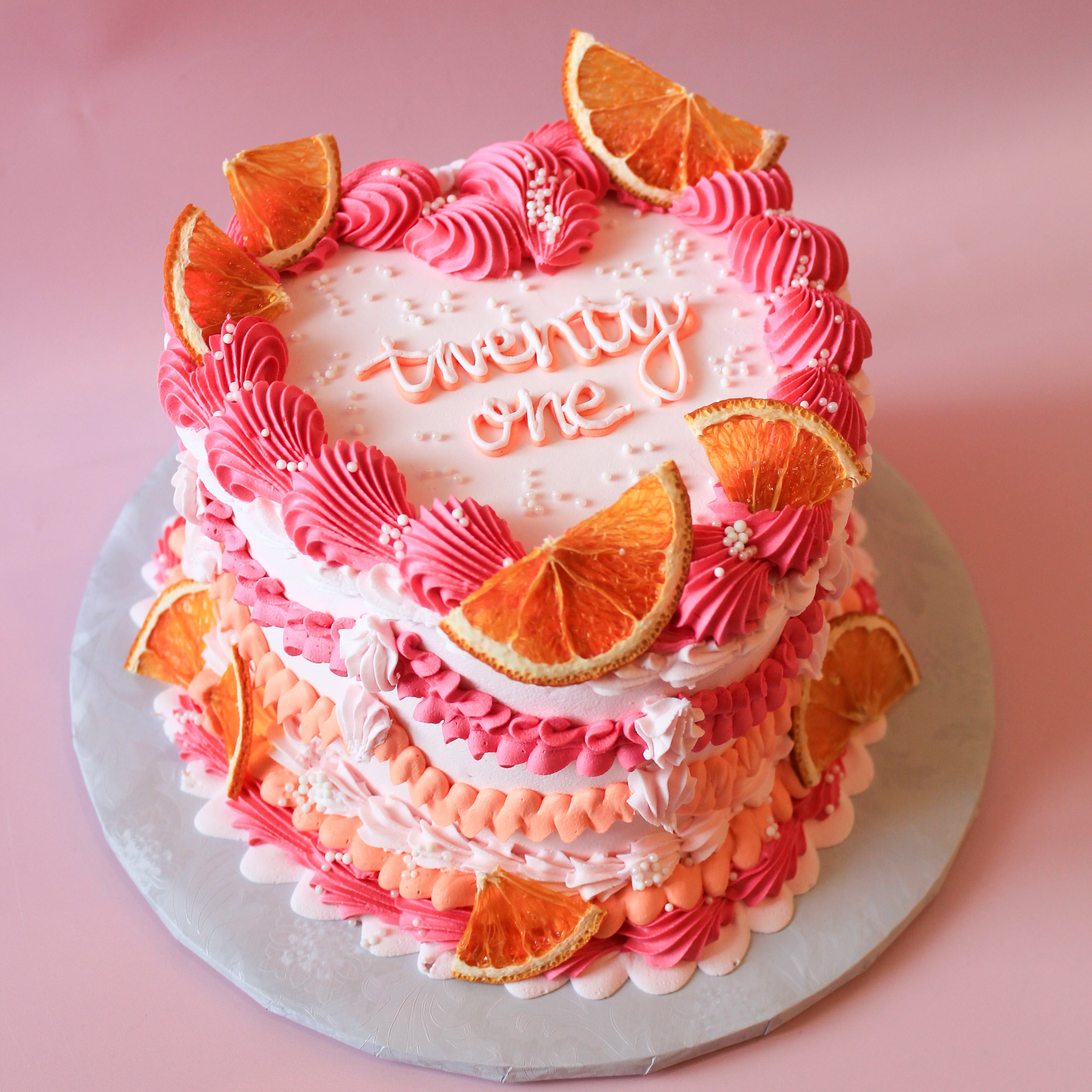 Two-tiered cake with pink and orange frosting, decorated with orange slices on a pink background.