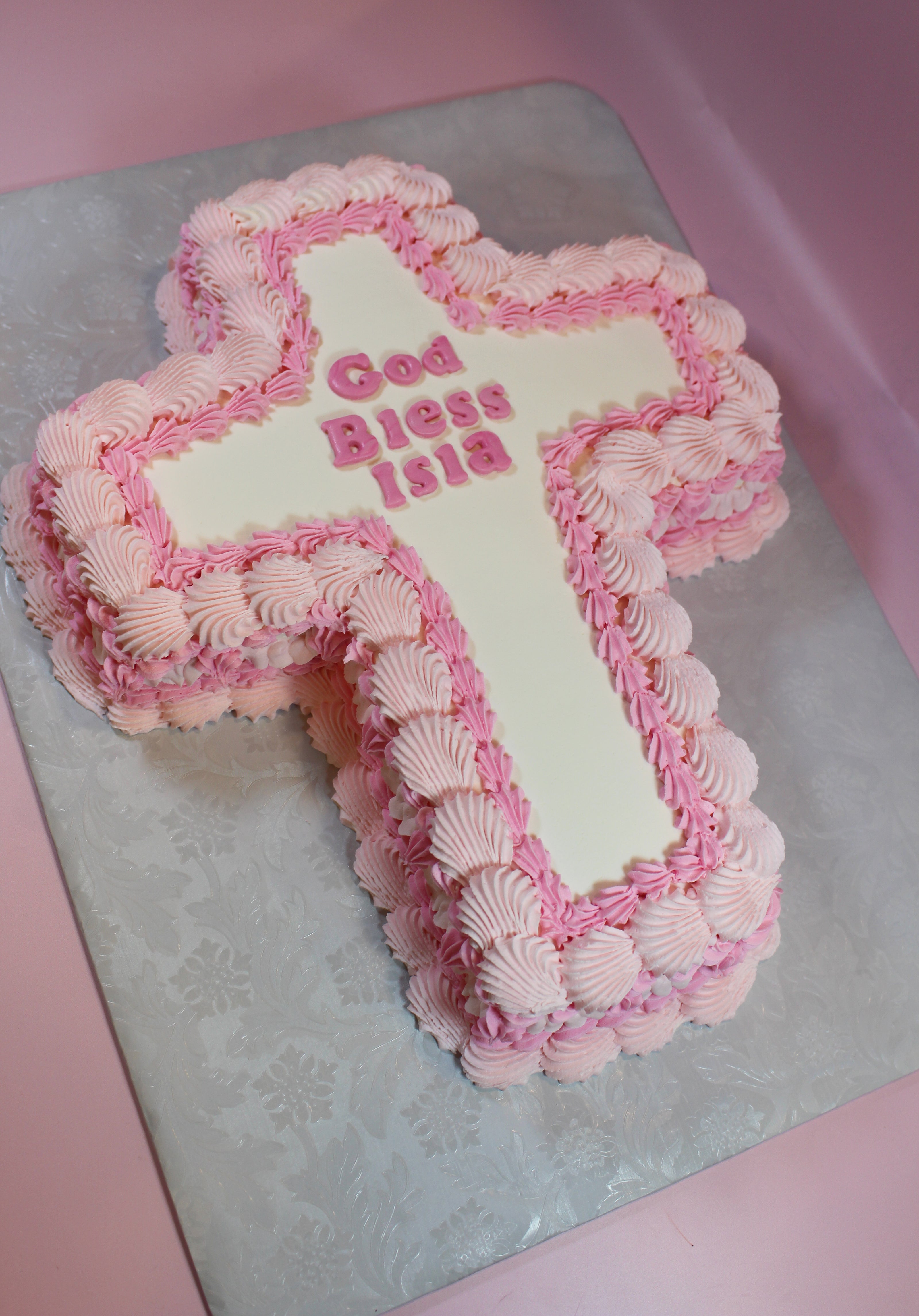 Cross-shaped cake with pink and white frosting on a silver cake board.