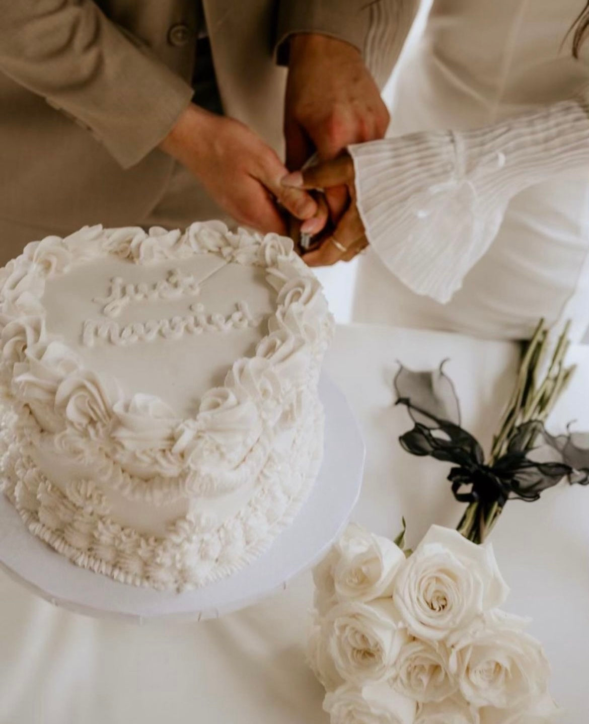Two people cutting a white wedding cake with a bouquet of flowers on a table.