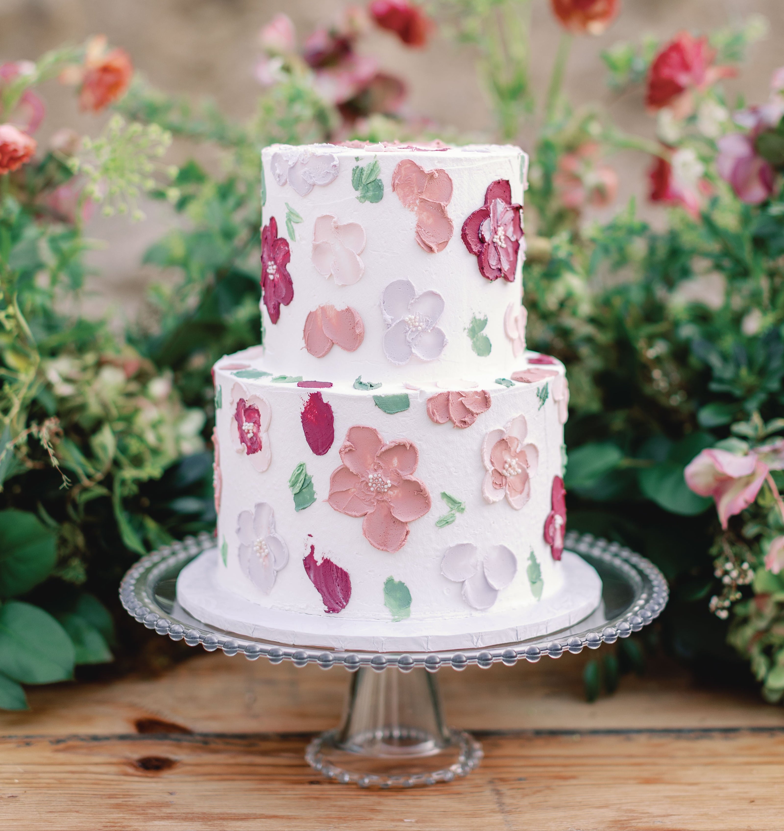 Two-tiered cake with floral design on a stand with flowers in the background